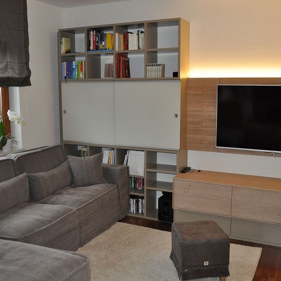 Ein modernes Wohnzimmer mit einer grauen Couch, einem Hocker, Teppich und einem hölzernen Entertainment-Schrank. Über der Couch befindet sich ein Bücherregal mit Büchern. Ein Flachbildfernseher ist an der Wand montiert.