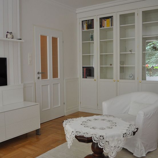 Ein Raum mit weißen Wänden und Möbeln, darunter ein weißer Stuhl mit einem Spitzentischdecken, ein Glasschrank und ein weißer Fernseher auf einem Regal.
