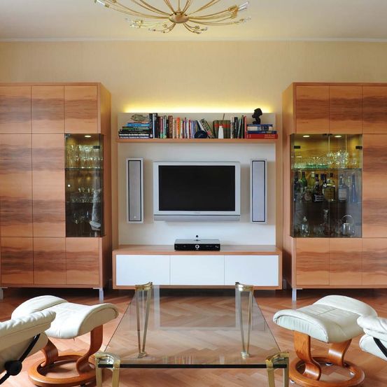 Ein modernes Wohnzimmer mit Holzmöbeln. Ein Glastisch steht in der Mitte, flankiert von zwei weißen Sesseln. Der Raum verfügt über einen an der Wand montierten Fernseher, eine Medienkonsole und Bücherregale auf beiden Seiten.
