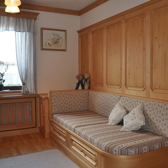 Ein gemütliches Zimmer mit Holzwänden und einer Bank unter einem Fenster. Die Bank hat einen blumenbedruckten Kissenbezug und zwei weiße Kissen. Ein Bilderrahmen und eine Puppe sind an der Wand. Ein Heizkörper befindet sich unter dem Fenster und eine Pflanze in einem Topf.