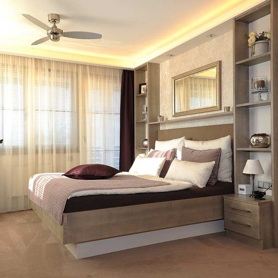 Ein modernes Schlafzimmer mit beigefarbenen Wänden, einem großen Bett mit Kissen, einem Spiegel und Regalen. Ein Deckenventilator und weiße Vorhänge schmücken den Raum.