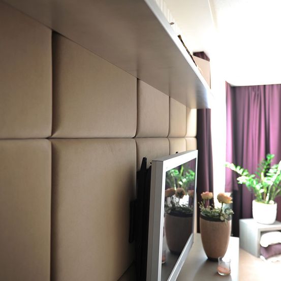Eine beige gepolsterte Wand mit einem montierten Fernseher reflektiert einen Blumenvase mit Pflanzen. Eine lila Gardine hängt auf der rechten Seite.
