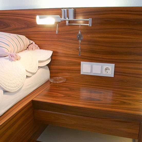 Ein Bett mit weißer Bettwäsche und einem Kissen auf einem hölzernen Kopfteil mit montierter Lampe und Steckdose.