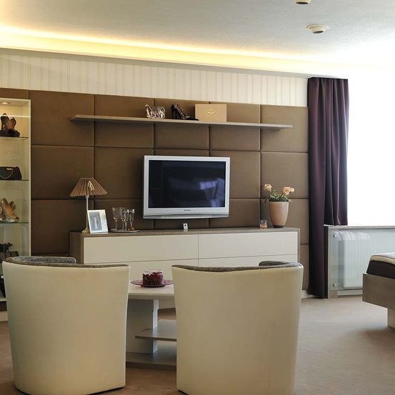 Ein modernes Wohnzimmer mit einem an der Wand montierten Fernseher, zwei weißen Stühlen und einem Tisch mit einer Lampe. Auf dem Regal befinden sich Gegenstände wie eine Box, High Heels und eine Lampe.