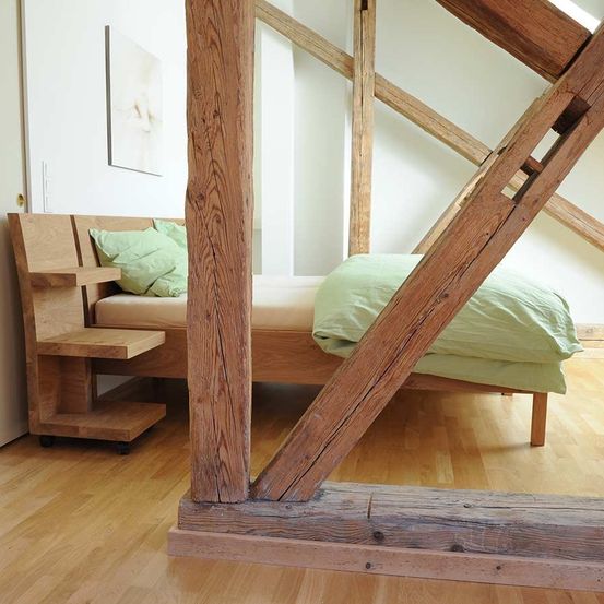 Ein Schlafzimmer mit Holzbettrahmen, Holzboden und freiliegenden Holzbalken. Das Bett hat eine beige Matratze, ein grünes Kissen und eine grüne Decke. Es gibt einen hölzernen Nachttisch mit Rädern und ein Gemälde an der Wand.