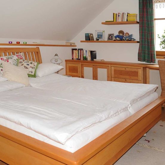 Ein ordentlich gemachtes Bett mit weißen Laken und Kissen befindet sich in einem gemütlichen Schlafzimmer. Zwei Holzregale an der Wand halten Bücher und dekorative Gegenstände. Ein Schreibtisch steht in der Nähe des Fensters mit einem grünen Vorhang.