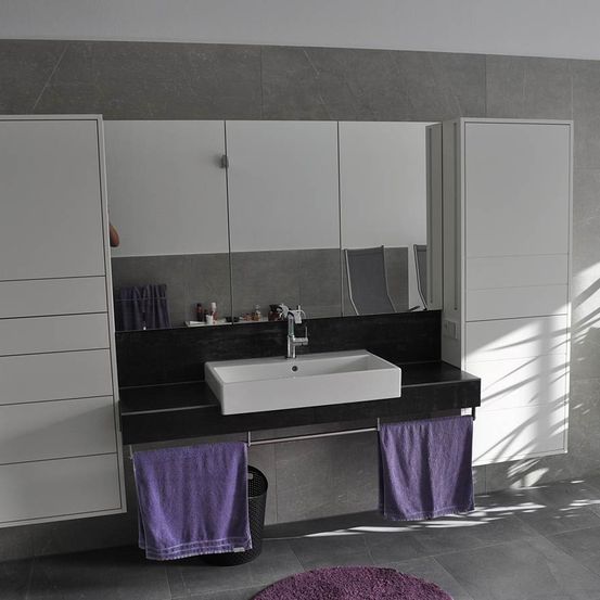 Ein modernes Badezimmer verfügt über ein weißes Waschbecken mit silberner Armatur, das auf einer schwarzen Arbeitsplatte steht. Der Raum ist mit grauen Fliesen und weißen Schränken ausgestattet. Lila Handtücher hängen an der Wand und ein runder lila Teppich liegt auf dem Boden.
