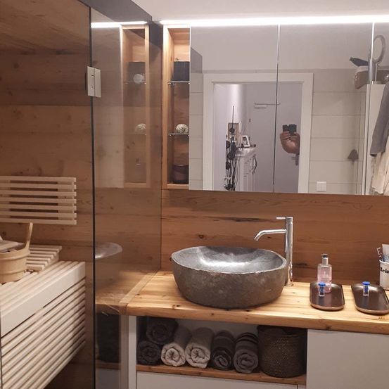 Ein Badezimmer mit Holzwand und -arbeitsplatte, ausgestattet mit einem Steinwaschbecken und einem Wasserhahn. Unter dem Waschbecken befinden sich gefaltete Handtücher und rechts zwei kleine Tabletts. Ein Spiegel reflektiert einen Raum mit einem Schrank und einer Tür.