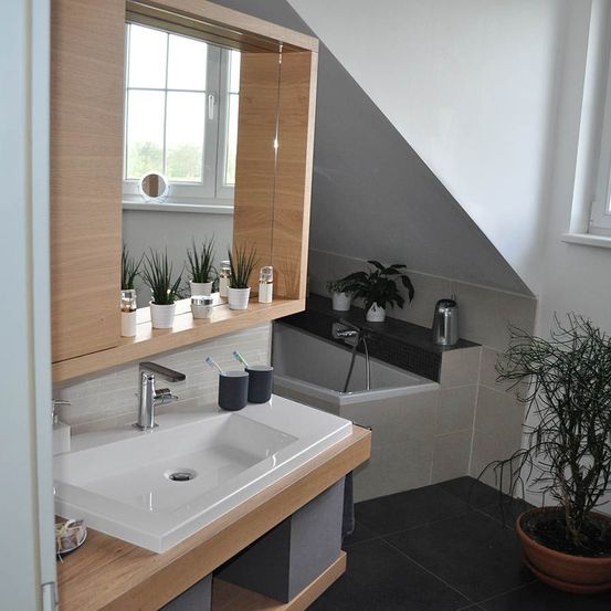 Ein modernes Badezimmer mit einem hölzernen Waschtisch, einem großen Spiegel, einem weißen Waschbecken und Topfpflanzen. Es gibt eine Badewanne mit einem Wasserhahn und einer Dusche in der Ecke. Die Wände sind weiß und der Boden ist mit schwarzen Fliesen belegt.