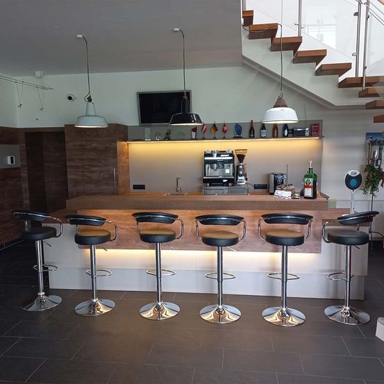 Eine moderne Küche mit Frühstücksbar, Kaffeemaschine und sechs schwarzen Barhockern. Der Raum hat Holzböden und eine Treppe.