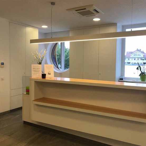 Der Empfangsbereich hat einen Schreibtisch mit einer Blumenvase und einem Topf. Es hat weiße Wände, eine Decke mit einem langen Licht und ein rundes Fenster. Vor dem Fenster steht ein Gebäude.