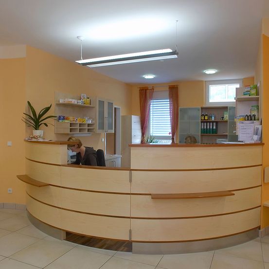 Ein Innenraum einer Rezeption in einer Klinik. Der Empfangstresen ist aus Holz mit einer geschwungenen Vorderseite. Eine Frau sitzt am Schreibtisch. Verschiedene Regale mit medizinischen Utensilien sind an der Wand montiert.