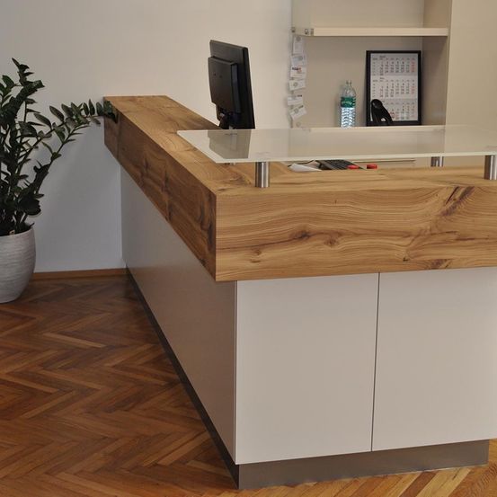 Eine moderne Rezeptionstheke mit Holzplatte und Glasfront, ausgestattet mit einem Computerbildschirm, Tastatur und einer Topfpflanze. Die Wand ist weiß, und auf einem Regal befinden sich ein Kalender und eine Wasserflasche.
