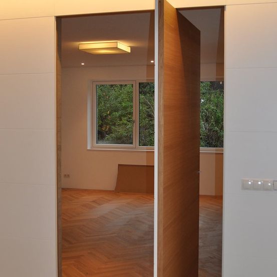Ein leerer Raum mit einer Holztür, einem offenen Fenster und einer Deckenleuchte. Der Raum hat Holzböden und weiße Wände.