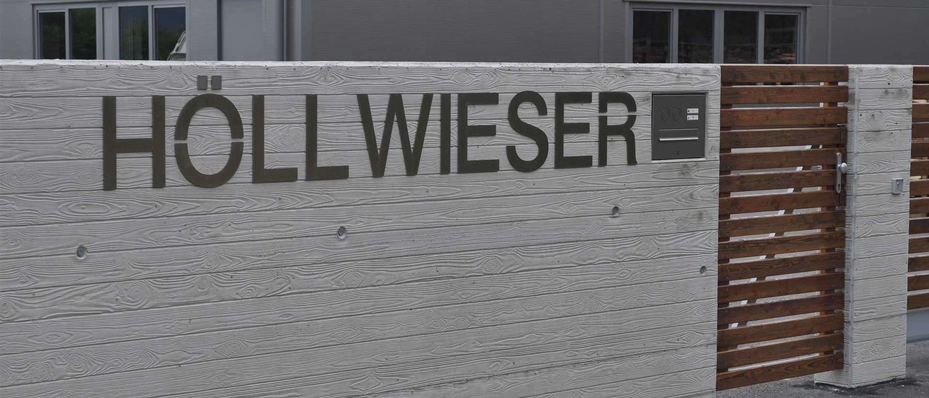 Ein hölzernes Schild mit dem Namen 'LL WIESER' in schwarz auf einem strukturierten grauen Hintergrund.