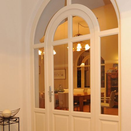 Eine gewölbte weiße Tür mit Glasscheiben spiegelt ein Esszimmer mit Kronleuchter, Tisch und Stühlen wider.