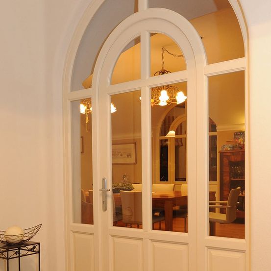 Eine gewölbte weiße Tür mit Glasscheiben spiegelt ein Esszimmer mit Kronleuchter, Tisch und Stühlen wider.