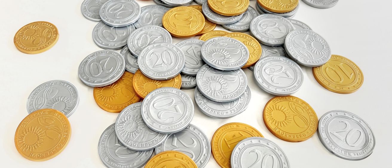 Ein Haufen Silber- und Goldmünzen mit verschiedenen Zahlen und Symbolen wie 10, 20 und 21 auf einer weißen Oberfläche angeordnet.