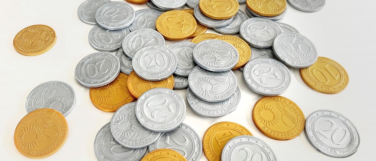 Ein Haufen Silber- und Goldmünzen mit verschiedenen Zahlen und Symbolen wie 10, 20 und 21 auf einer weißen Oberfläche angeordnet.