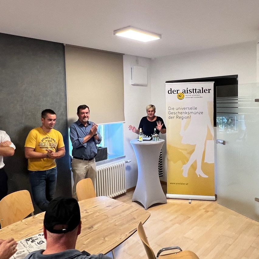 Eine Gruppe von Menschen steht in einem Raum mit einem Banner, das 'der aistaler' steht. Eine Frau steht hinter einem Tisch mit Flaschen und klatscht.