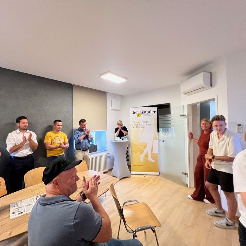 Eine Gruppe von Menschen in einem Raum, einige klatschen, eine Person spricht an einem Podium. Ein Banner mit 'der aistaler' ist sichtbar.