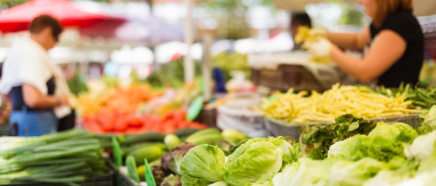 Ein geschäftiger Markt voller verschiedener frischer Gemüsesorten wie Tomaten, Salat, Gurken und Paprika. Einige sind unter roten Sonnenschirmen.