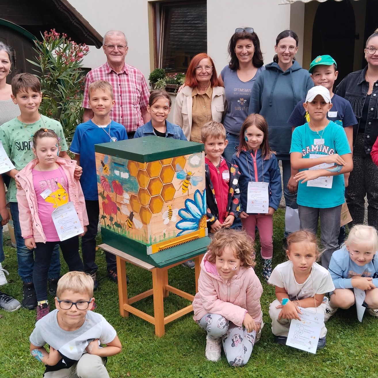 Eine Gruppe von Kindern und Erwachsenen posiert für ein Foto vor einem Bienenstock, der mit Bienen und Blumen bemalt ist.