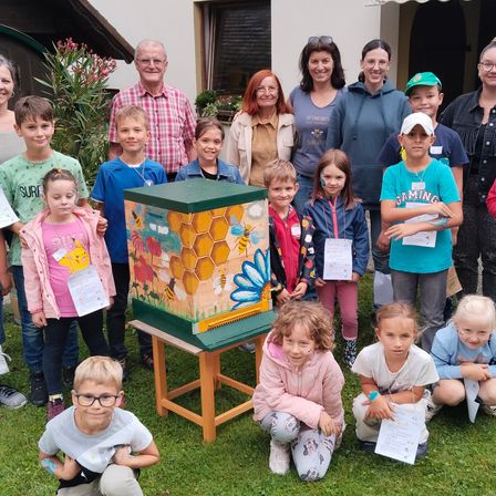 Eine Gruppe von Kindern und Erwachsenen posiert für ein Foto vor einem Bienenstock, der mit Bienen und Blumen bemalt ist.