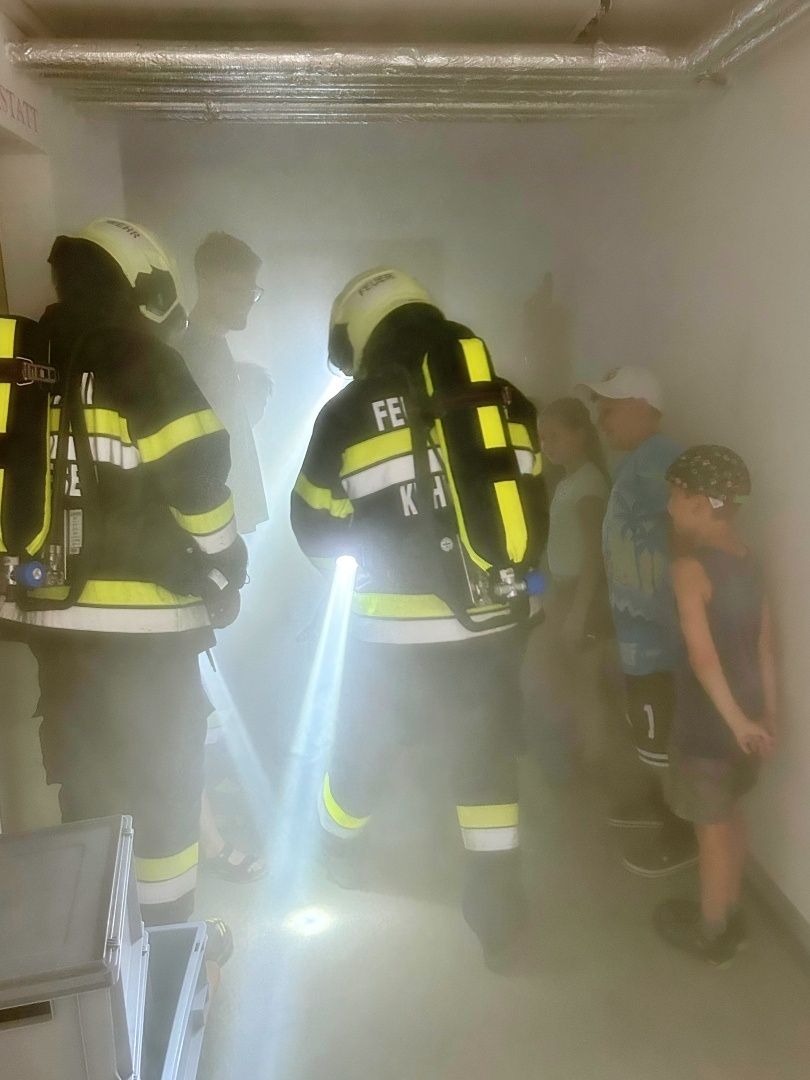 Zwei Feuerwehrleute in voller Montur navigieren durch einen verrauchten Korridor, einer hält eine Taschenlampe. Sie werden von mehreren Zuschauern begleitet, darunter ein junges Kind und zwei Erwachsene.