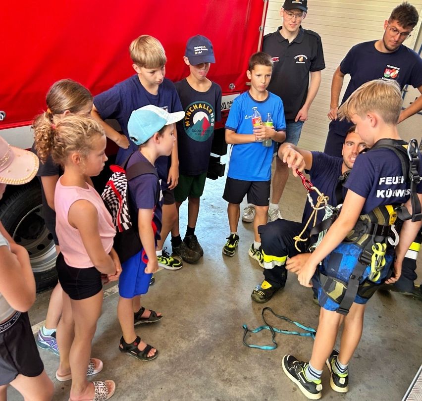 Eine Gruppe von Kindern lernt von einem Mann in einer Feuerwehruniform über das Löschen von Bränden. Sie stehen in einer Garage, mit einem Feuerwehrwagen im Hintergrund.