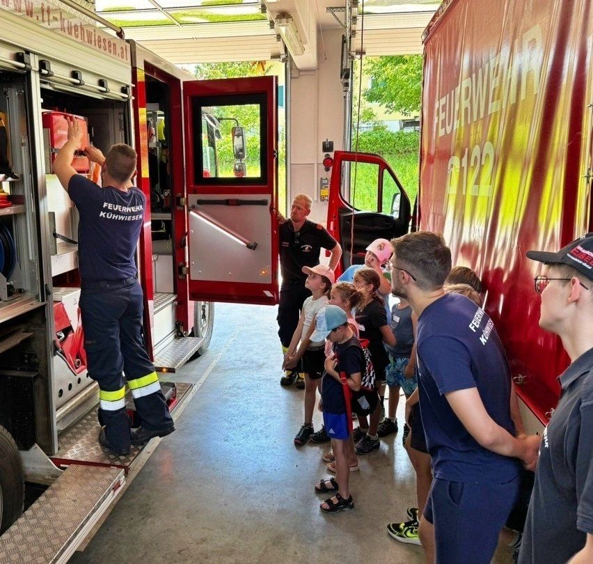 Feuerwehrleute zeigen einer Gruppe von Kindern das Innere eines Feuerwehrwagens. Sie erklären die Ausrüstung und Werkzeuge, die von den Feuerwehrleuten verwendet werden.