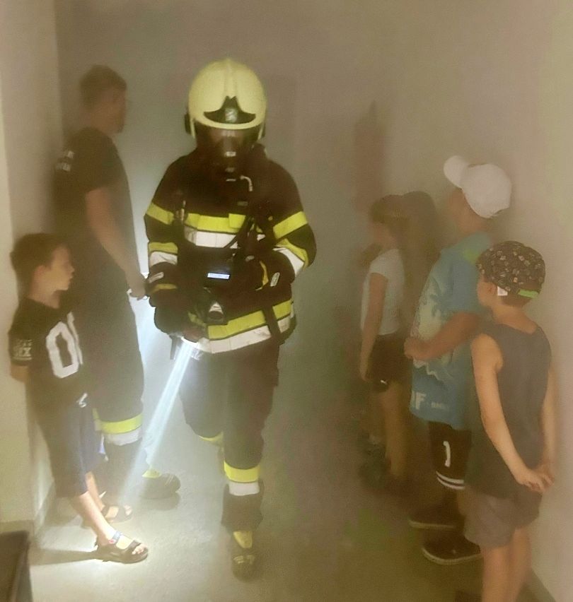 Ein Feuerwehrmann in voller Ausrüstung geht mit einer Taschenlampe durch einen rauchigen Flur, während mehrere Kinder und Erwachsene beobachten.