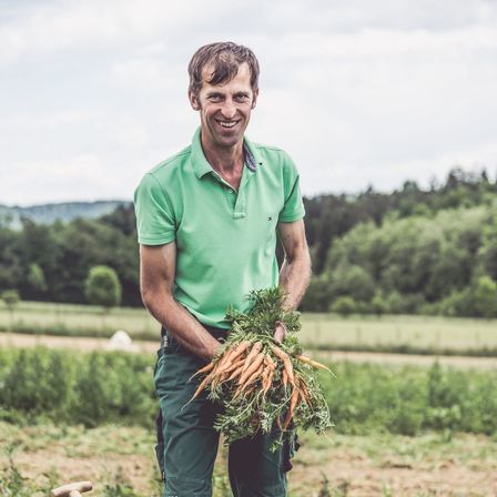 Ein Mann in einem grünen Poloshirt und grüner Hose hält einen Bund Karotten auf einem Feld.
