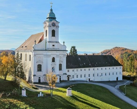 Eine große weiße Kirche mit grünem Turm und einer Uhr steht vor einem grasigen Feld mit Bäumen. Eine lange Auffahrt führt zu dem Gebäude.