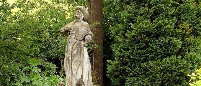 Eine Statue einer Frau im Garten, die einen Hut trägt und ein Buch hält. Die Statue steht auf einem Podest mit Kinderstatuen an der Basis. Der Garten ist von verschiedenen Pflanzen und Bäumen umgeben.