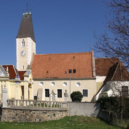 Eine alte Kirche mit Glockenturm, rotem Dach und mehreren Fenstern. Sie steht auf einer Steinmauer mit einem Rasenbereich davor.
