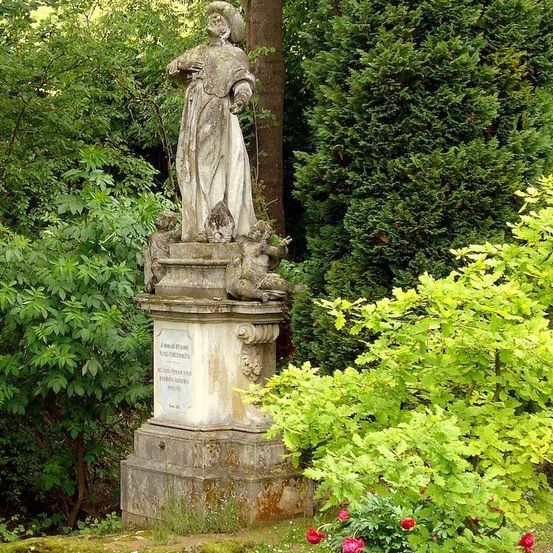 Eine Statue einer Person mit Hut und offenem Buch steht auf einem Podest in einem Garten mit verschiedenen Pflanzen und Bäumen.