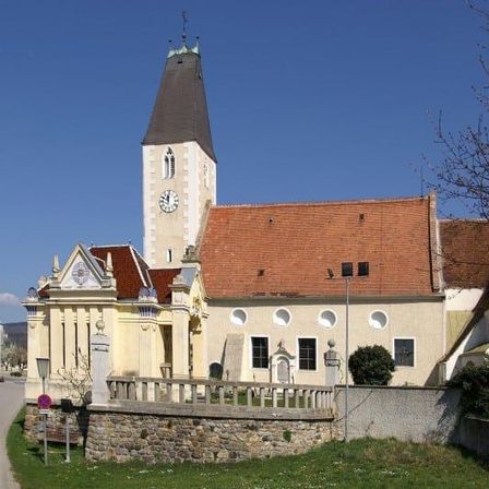 Eine historische Kirche mit einem hohen Turm und einer Uhr, vor dem Hintergrund eines klaren blauen Himmels. Das Gebäude hat ein rotes Dach und ist von einer Steinmauer und einem kleinen Garten umgeben.