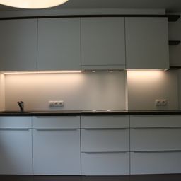 Eine moderne Küche mit weißen Schränken, schwarzen Arbeitsplatten und eingebauter Beleuchtung. Es gibt ein Spülbecken mit einem Wasserhahn und einen Herd mit Dunstabzugshaube.