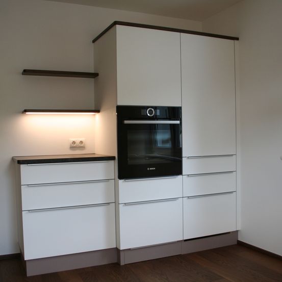Eine moderne Küche mit weißen Schränken, einem schwarzen Ofen und einer dunklen Arbeitsplatte. Über dem Ofen befindet sich ein kleines Regal mit einer Lampe.