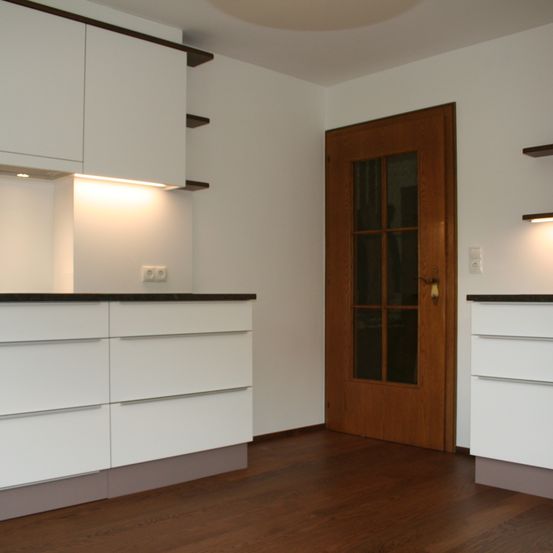 Eine moderne Küche mit weißen Schränken, einer Holztür und einem Holzboden. Die Wände sind weiß, und es gibt Einbauleuchten über den Schränken.