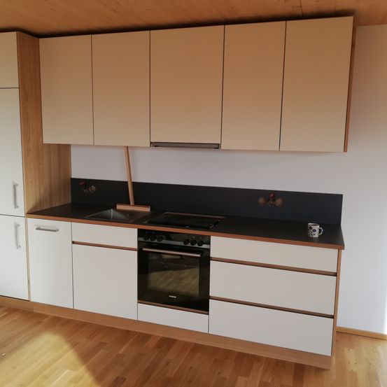 Eine moderne Küche mit Holzboden, weißen Schränken und einer schwarzen Arbeitsplatte. Ein Ofen und ein Waschbecken befinden sich zentral, mit einer Kaffeetasse auf der Arbeitsplatte.
