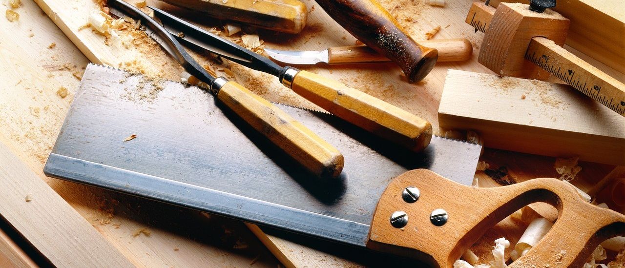 Eine Nahaufnahme verschiedener Holzbearbeitungswerkzeuge auf einem Holzarbeitstisch. Zu den Werkzeugen gehören Meißel, ein Hammer und eine Handsäge. Holzspäne sind auf der Oberfläche verstreut.