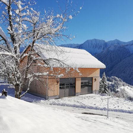 Ein Holzchalet mit schneebedecktem Dach steht in einer verschneiten Landschaft, umgeben von Bergen. Ein Baum mit schneebedeckten Ästen steht neben ihm.