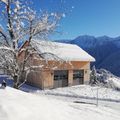 Ein Holzchalet mit schneebedecktem Dach steht in einer verschneiten Landschaft, umgeben von Bergen. Ein Baum mit schneebedeckten Ästen steht neben ihm.