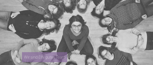 Eine Gruppe lächelnder Frauen, die in einem Kreis angeordnet sind, eine Frau in der Mitte mit Brille und Uhr. Das Bild hat einen Schwarz-Weiß-Filter.