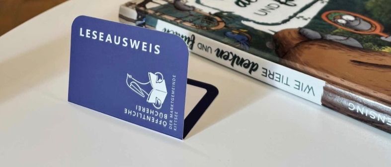 Ein Buch mit dem Titel 'Wie Tiere denken und fühlen' liegt neben einem blauen Lesezeichen mit 'LESEAUSWEIS' und einem Logo auf einem weißen Tisch, mit einem Bücherregal im Hintergrund.