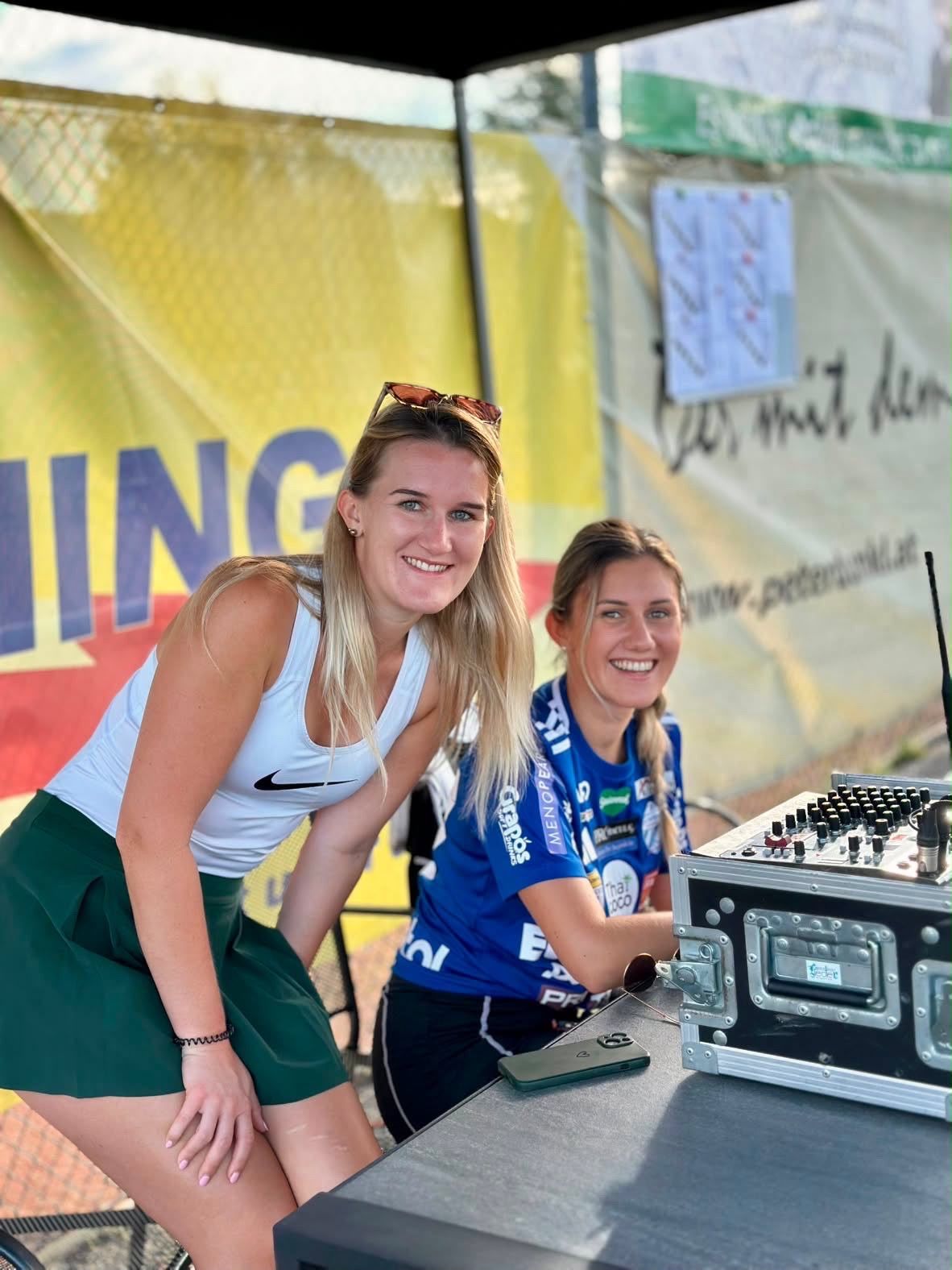 Zwei Frauen lächeln und posieren für ein Foto vor einem DJ-Pult, eine trägt ein weißes Tanktop und einen grünen Rock, die andere ein blaues Shirt mit Mikrofon.