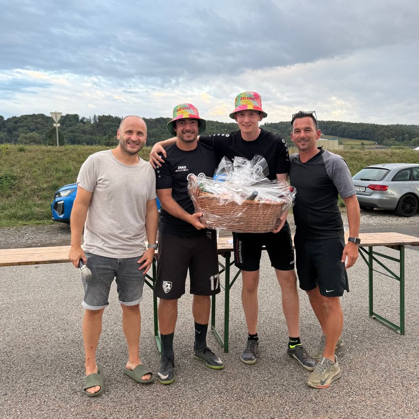 Vier Männer stehen auf einem Parkplatz, zwei halten einen Korb mit einem Geschenk darin, während die anderen lächeln und für ein Foto posieren.