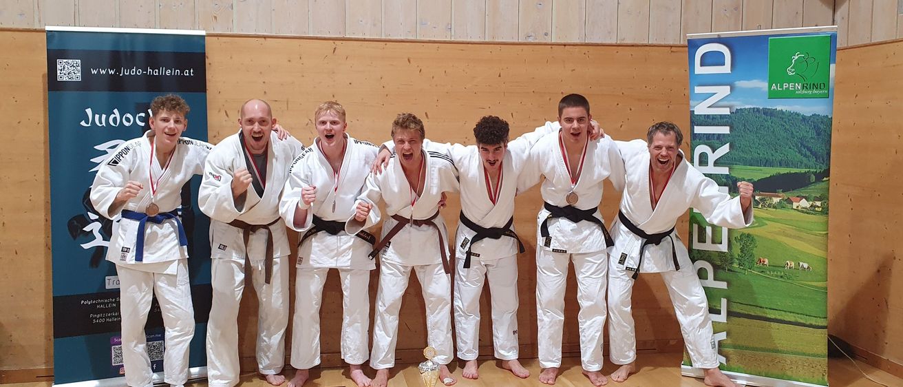 Sechs Männer in weißen Karate-Uniformen stehen nebeneinander, lächeln und posieren für ein Foto. Sie haben Medaillen um den Hals. Sie stehen vor einer Holzwand.